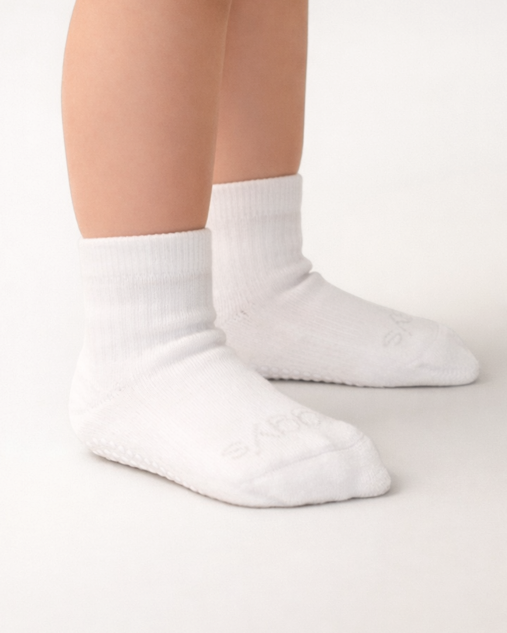 Toddler with a light skin tone wearing a pair of white toddler crew socks on an off-white background.