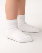 Toddler with a light skin tone wearing a pair of white toddler crew socks on an off-white background.