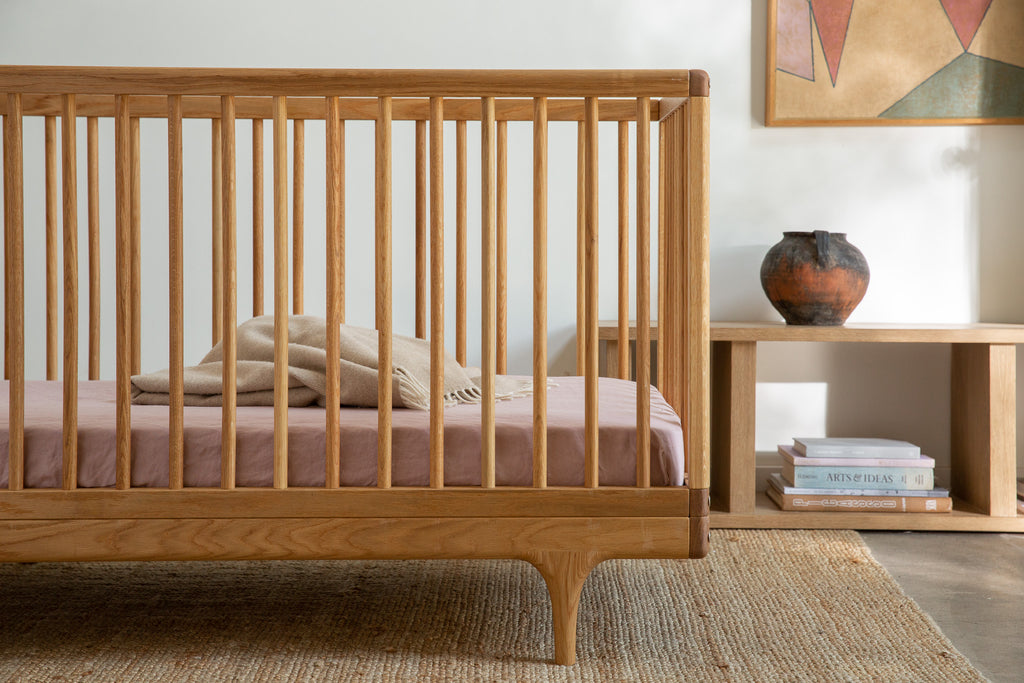 Caravan Crib in white oak with dusty pink fitted sheet and beige fringed throw, art books and ceramic vase staged nearby.
