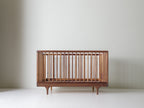 Caravan Crib in walnut, three-quarter view on a white studio floor, with lighter sapwood spindles against the dark frame.