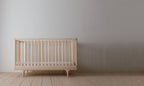 Caravan Crib in ash, front view on a maple plank floor, vertical turned spindles and tapered legs at each end.
