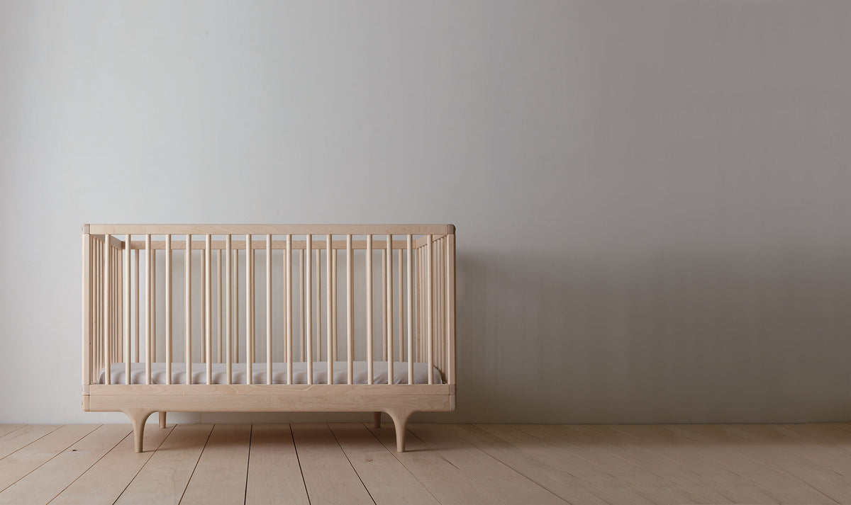 Caravan Crib in ash, front view on a maple plank floor, vertical turned spindles and tapered legs at each end.