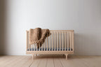 Caravan Crib in ash on a maple plank floor, chunky-knit caramel wool blanket draped over the side rail.