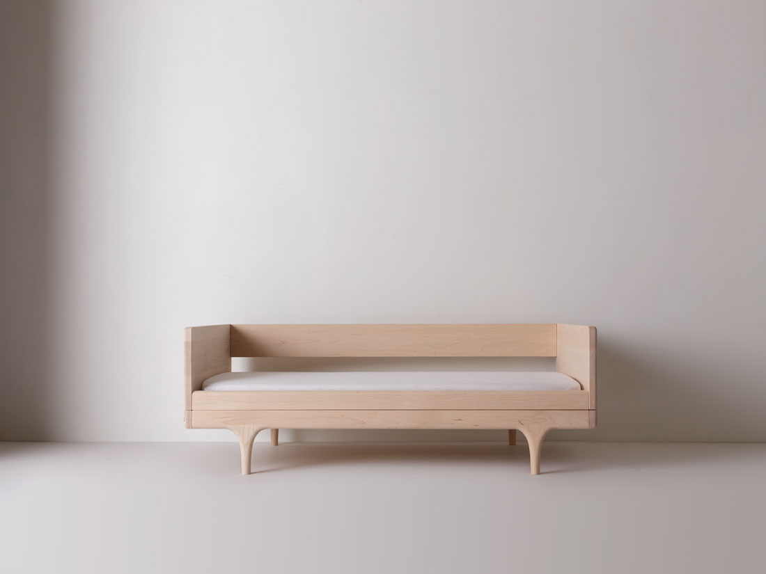 Caravan Divan in ash with backrest, low daybed configuration on a white studio floor, sculpted legs and a slim mattress.