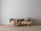 Caravan Divan in ash with backrest on a maple plank floor, chunky-knit caramel blanket draped across, sage cushion at right.