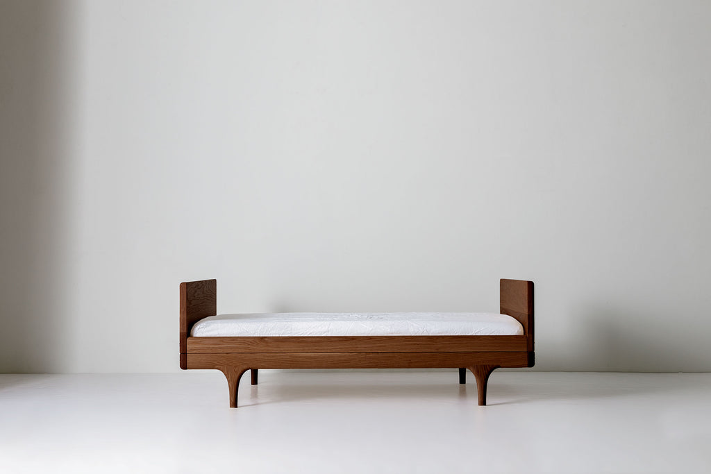 Caravan Divan in solid walnut, no-backrest configuration, mattress on the base on a white studio floor with sculpted legs.