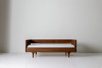 Caravan Divan in solid walnut with backrest, front view on a white studio floor, mattress on the base above sculpted legs.