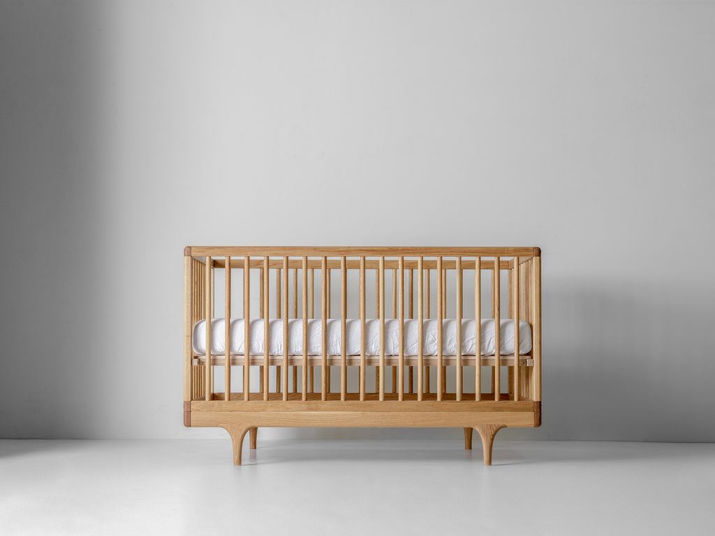 Caravan Crib in white oak with walnut corner caps, front view, mattress at higher support height on a white floor.