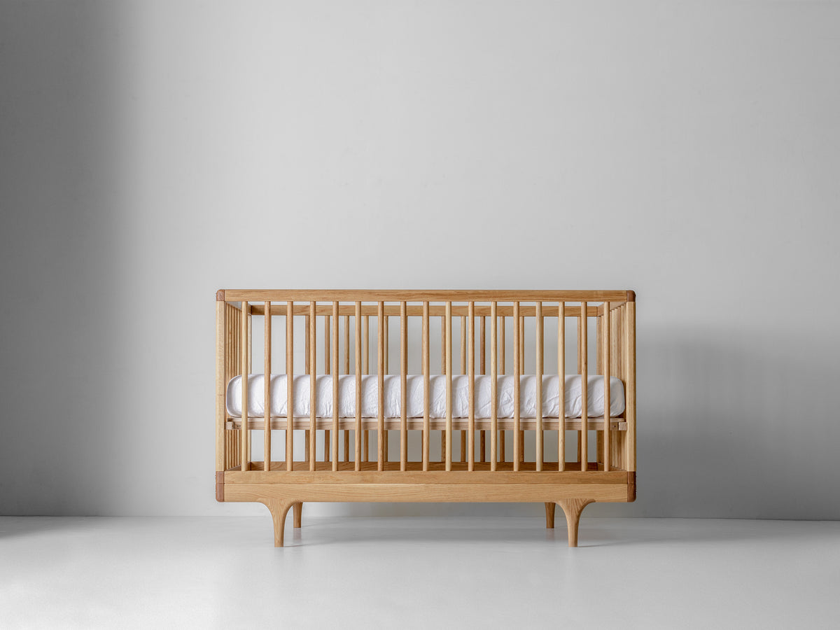 Caravan Crib in white oak with walnut corner caps, front view, mattress at higher support height on a white floor.
