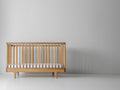Caravan Crib in white oak with walnut corner caps, front view on a white studio floor, mattress at lowest height.