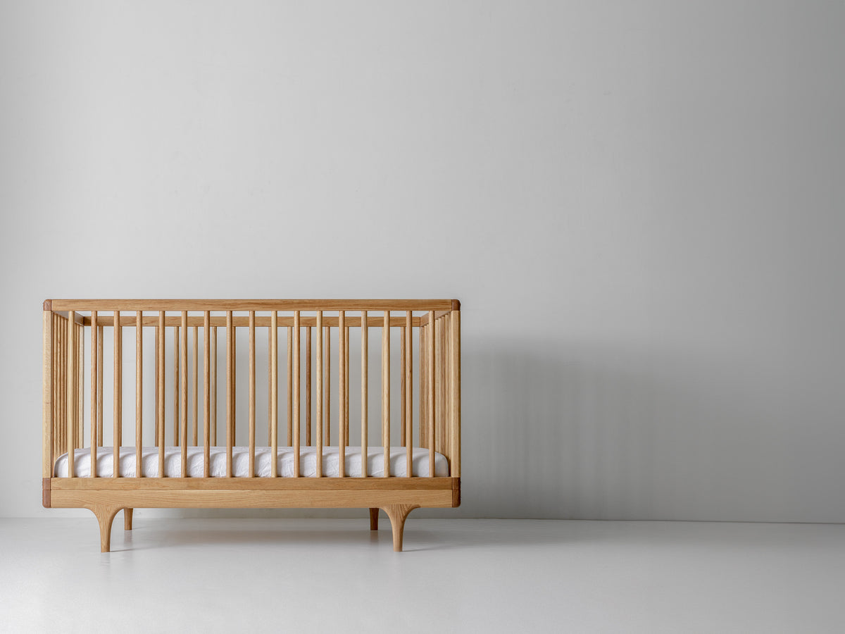 Caravan Crib in white oak with walnut corner caps, front view on a white studio floor, mattress at lowest height.