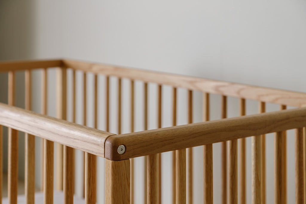 Caravan Crib in white oak, top corner detail showing the rounded rail meeting the post with a visible hex bolt.