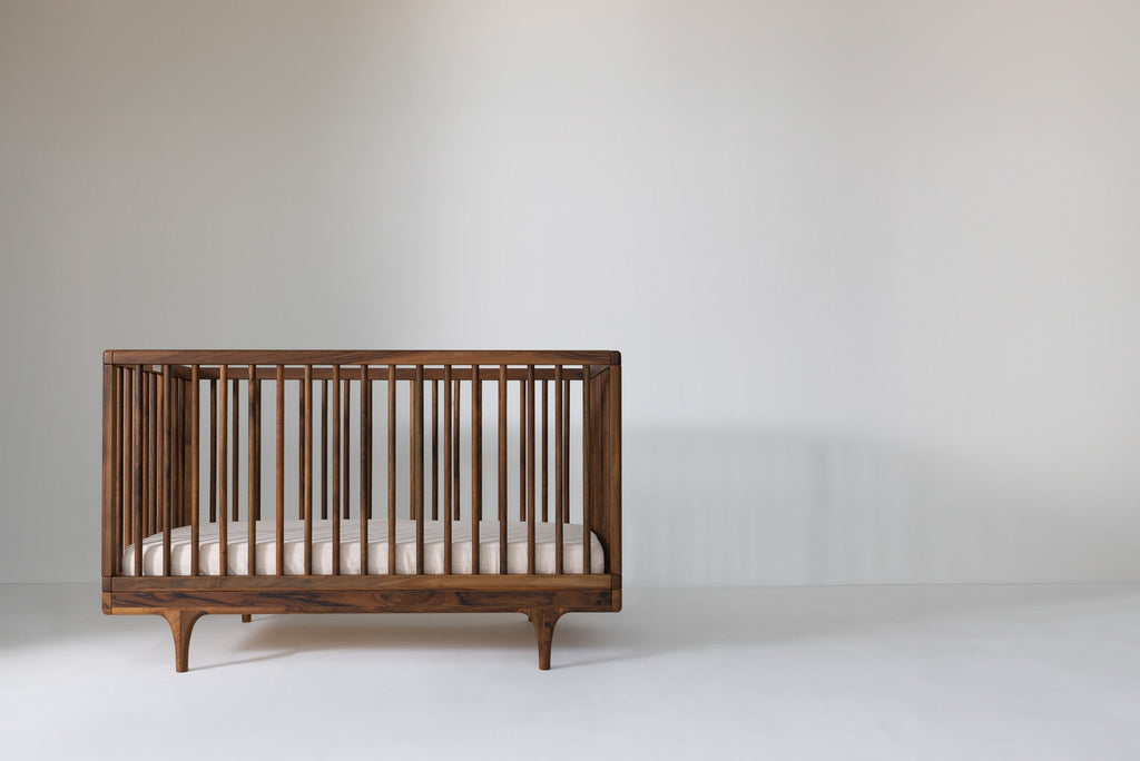 Caravan Crib in solid walnut, front view off-center to the left on a white floor, mattress at lower support height.