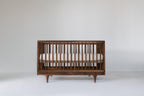 Caravan Crib in solid walnut, front view, mattress at the higher support height for newborns on a white studio floor.
