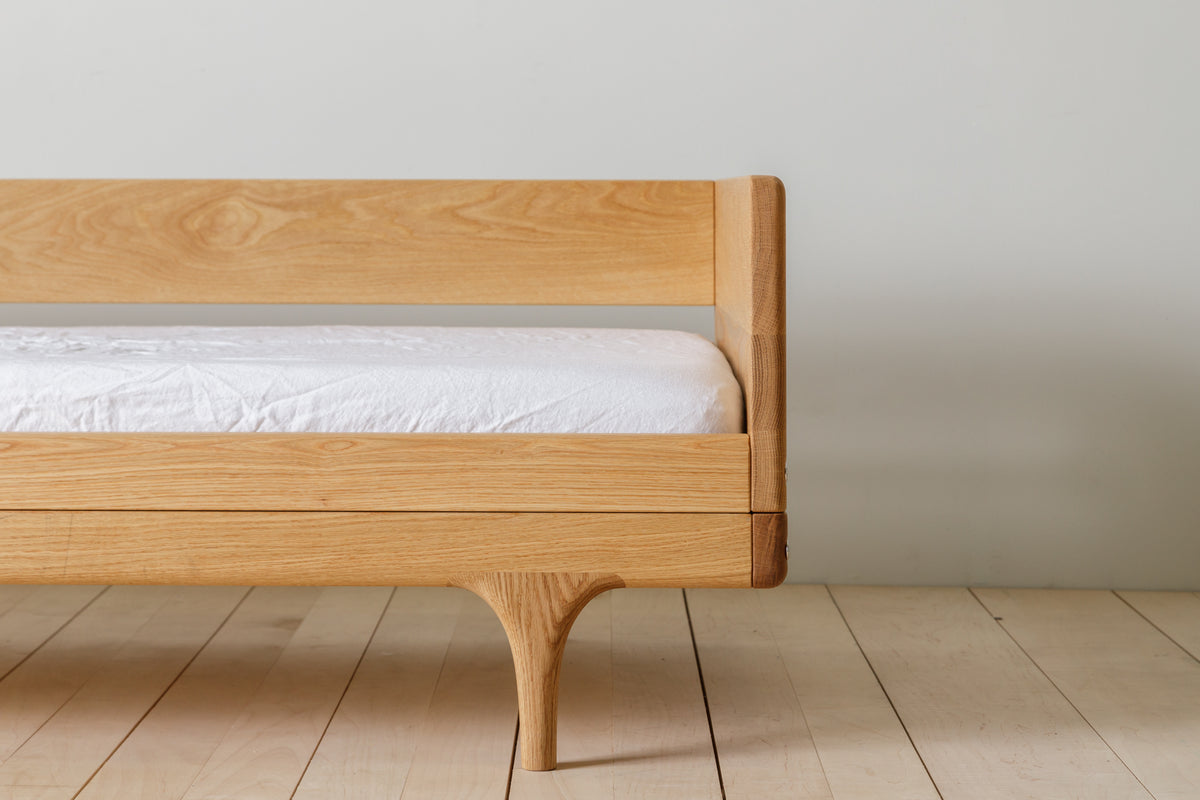 Caravan Divan in white oak, side close-up with backrest, sculpted leg, walnut corner cap on a maple plank floor.