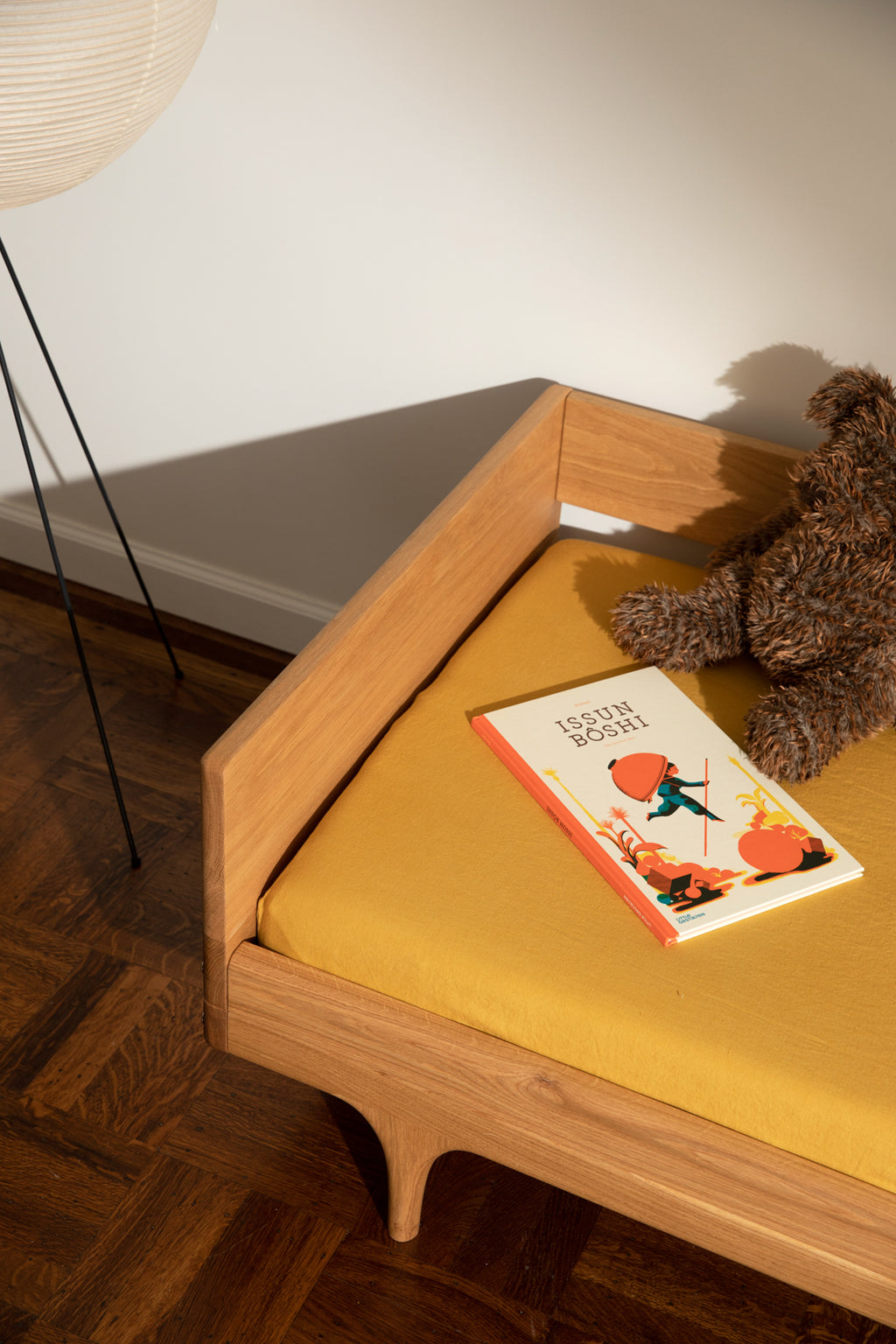 Caravan Divan in white oak with mustard sheet, Issun Bōshi book, and brown plush teddy, parquet floor with black floor lamp.