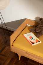 Caravan Divan in white oak with mustard sheet, Issun Bōshi book, and brown plush teddy, parquet floor with black floor lamp.