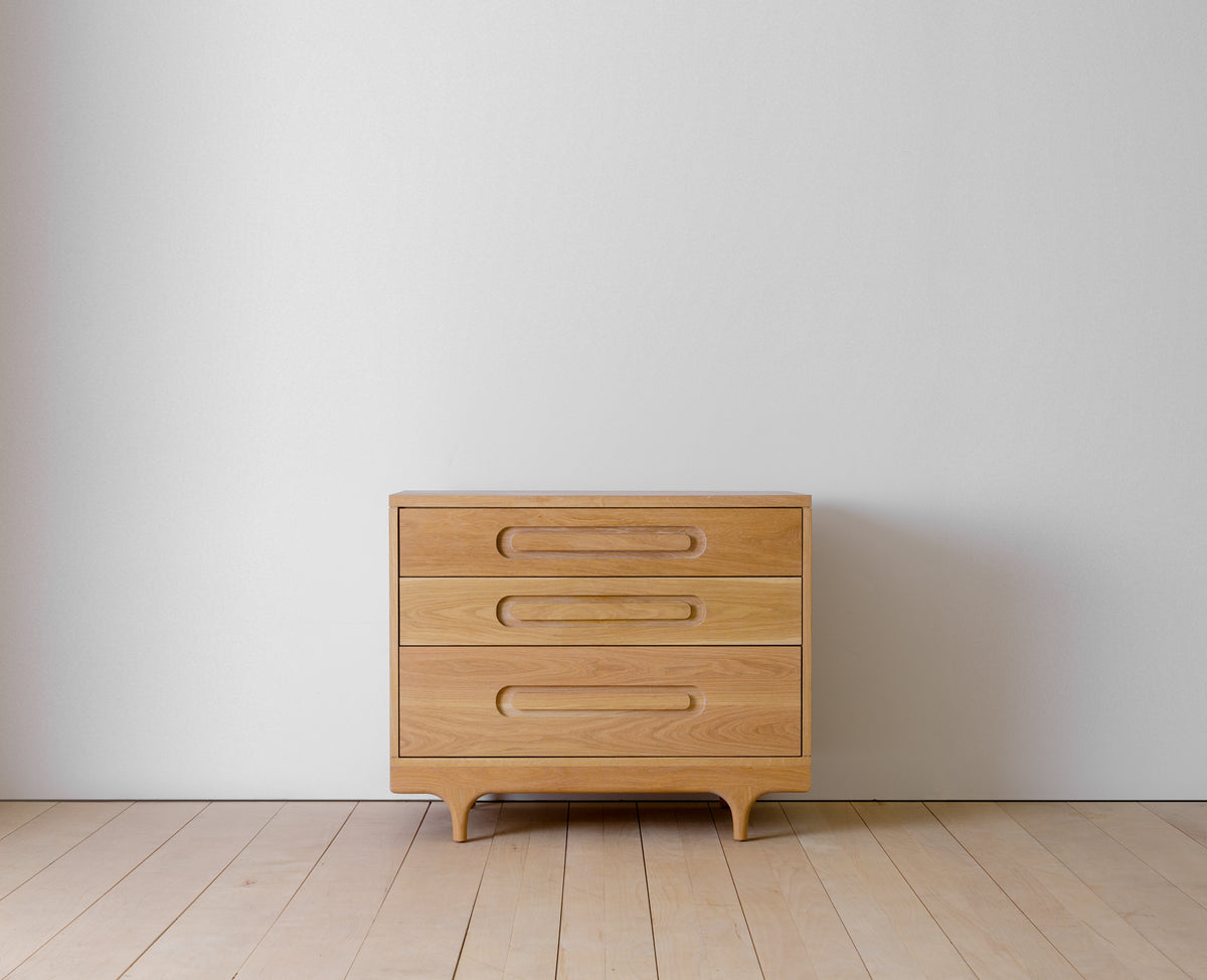 Caravan Dresser in white oak, front view on a maple plank floor against a pale grey wall, three drawers and sculpted legs.