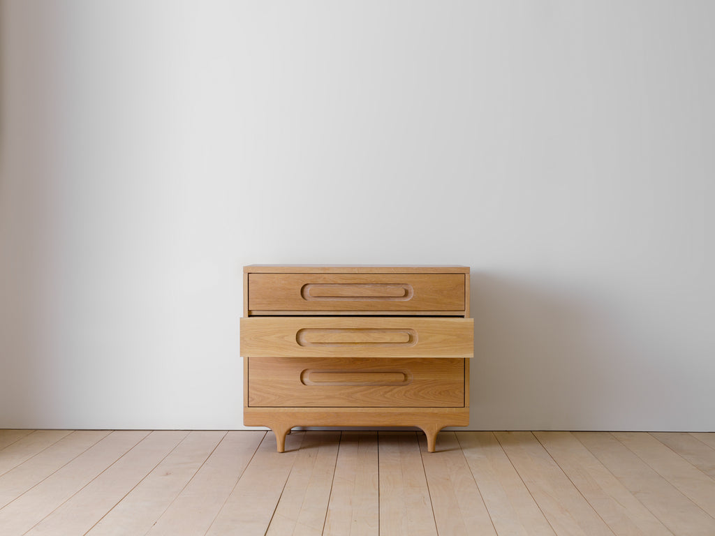 Caravan Dresser in white oak with the middle drawer pulled out, front view on a maple plank floor against a grey wall.