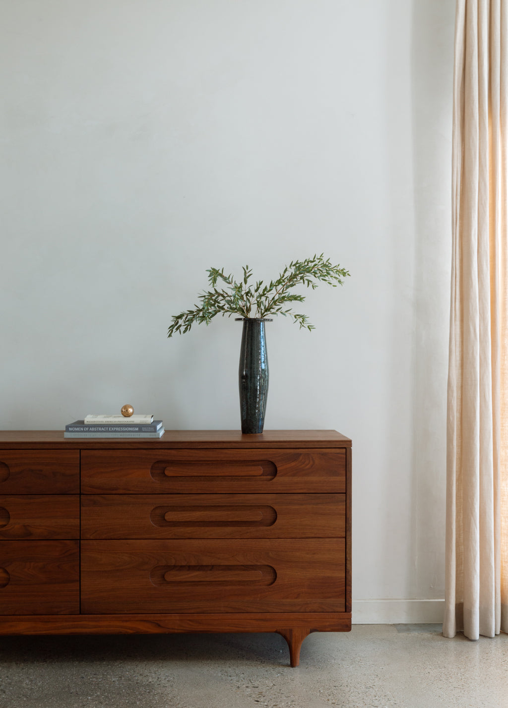 Caravan Double Dresser in walnut with tall dark iridescent vase of olive branches, art-book stack with copper sphere on top.