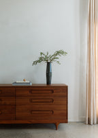 Caravan Double Dresser in walnut with tall dark iridescent vase of olive branches, art-book stack with copper sphere on top.
