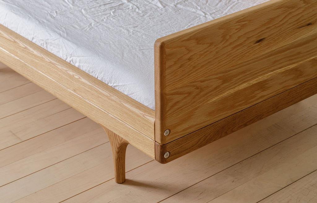 Caravan Divan in white oak, footboard corner detail with figured grain, sculpted leg, hex bolts, and walnut corner caps.