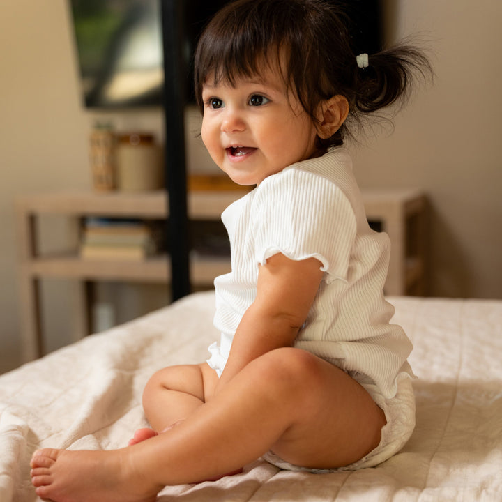 Smiling baby in a Dyper diaper and white top sitting on a bed