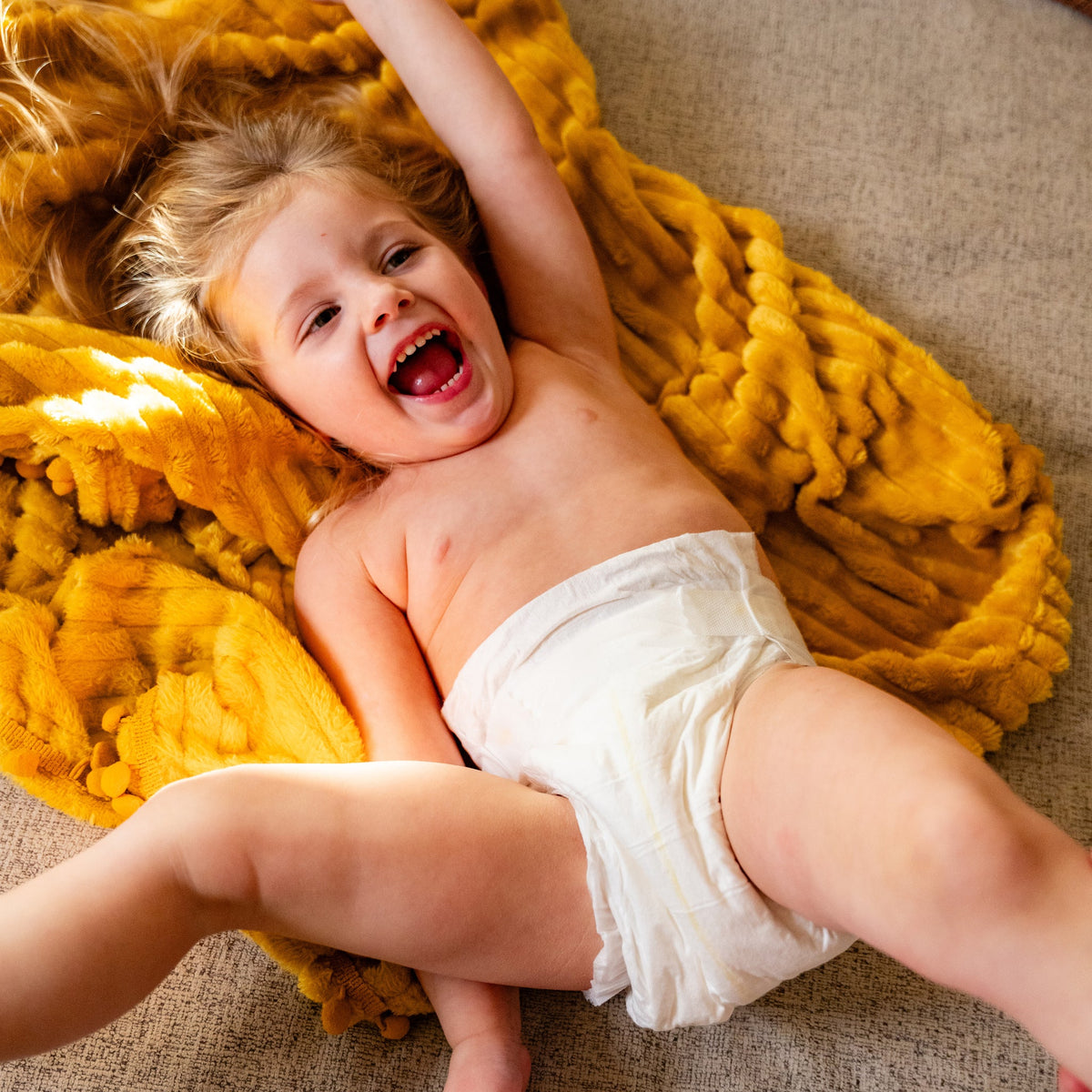 Laughing toddler in a Dyper diaper lying on a mustard knit blanket