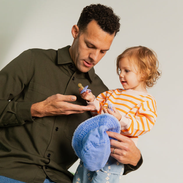 Father offering a BIBS pacifier to a toddler in a striped top