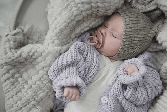 Mushie — A baby wearing a knitted hat and a lavender sweater sleeps peacefully, holding a pacifier in their m