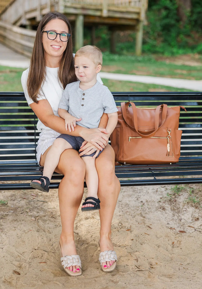 Baldwin Baby Company — Woman with glasses holding a young boy on a park bench, brown diaper bag beside them.