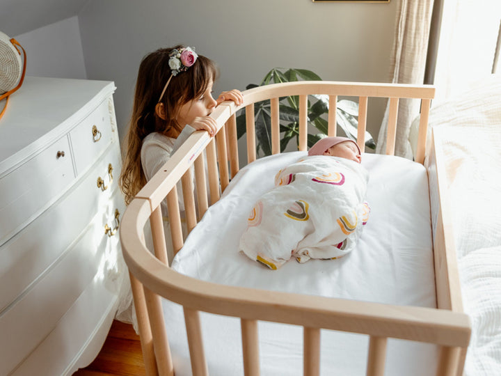Girl looking at a baby asleep in a babybay bedside co-sleeper