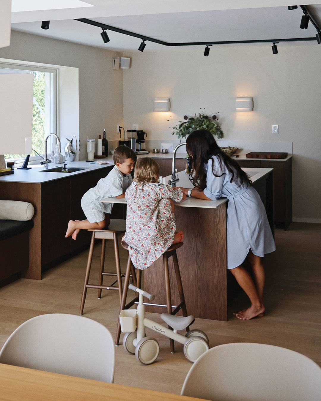 Mushie — Woman and two children in a modern kitchen with a tricycle on the floor.