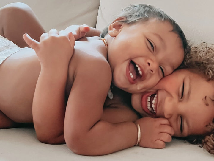 Two laughing toddlers cuddling on a sofa, one in a Parasol diaper